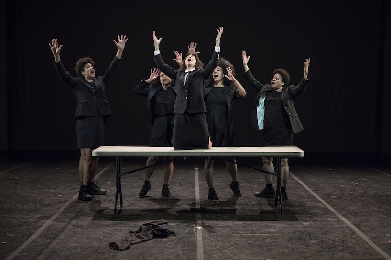 Four women sing around a table with their arms outstretched. Another woman knees atop the table and sings as well.